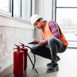 handsome fireman in helmet sitting near extinguishers and holding clipboard